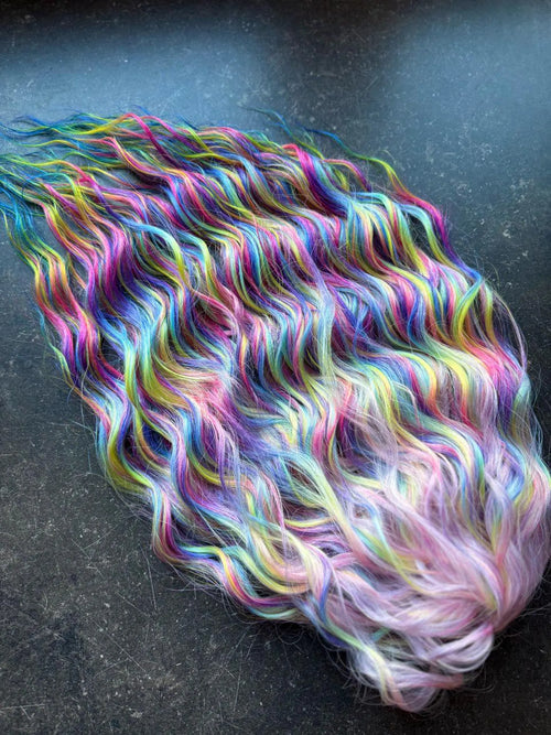 Wavy rainbow ombre hair extensions in pink, blue, purple, and yellow on dark background.