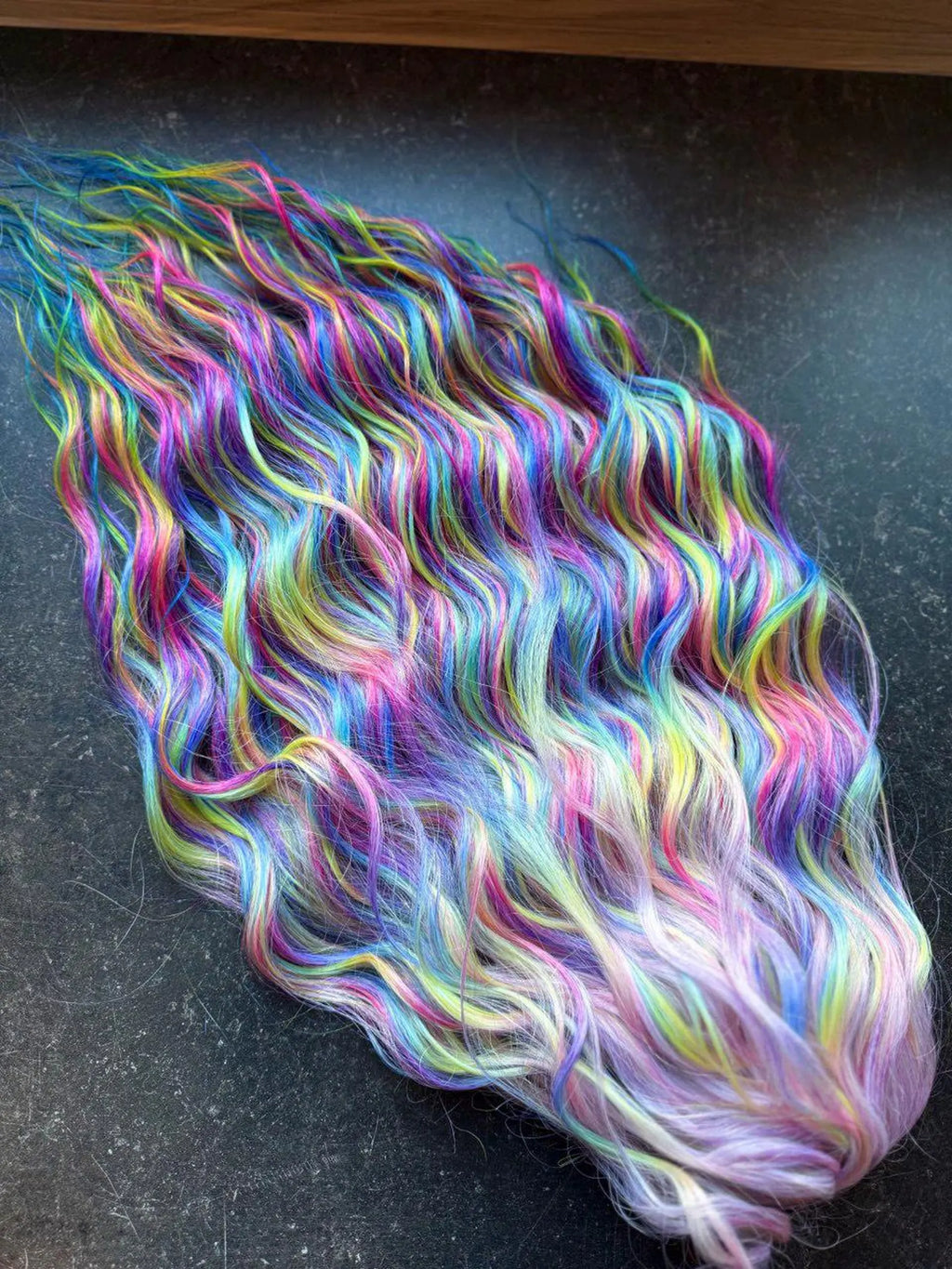 Wavy rainbow ombre hair extensions in pink, blue, purple, yellow, green on dark surface.